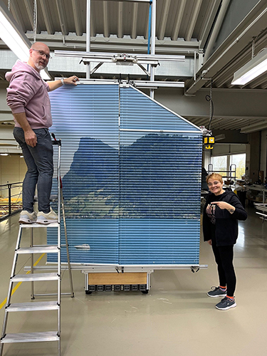 zwei aufgespannte Plissees mit einem Mann auf einer Leiter davor und einer Frau rechts daneben stehend